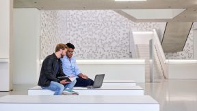 Zwei Studierende sitzen vor einem Laptop im Foyer der Universität.